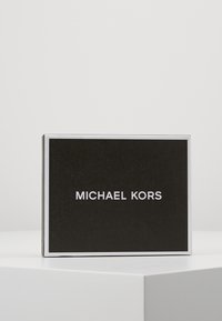 Boîte noire avec une texture, affichant le texte « MICHAEL KORS » en blanc audacieux. Bords soulignés par une fine bordure argentée.