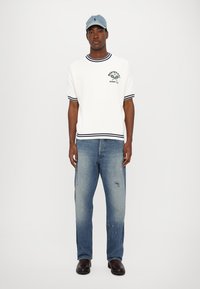 Hvit t-skjorte med korte ermer og marineblå striper, med en logo, kombinert med falmede blå jeans og svarte boots. En denimcap fullfører stilen.