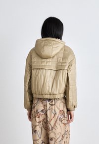 Khaki puffer jacket with a hood, featuring quilted texture and elastic hem. Paired with patterned beige trousers showcasing equestrian motifs.