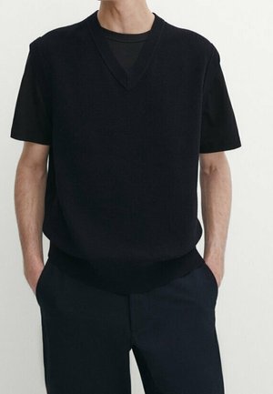 Homme portant un pull noir à manches courtes en V sur un t-shirt noir à col rond avec un pantalon noir, les mains dans les poches, sur un fond uni.