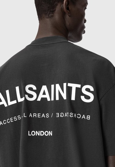 Ман, носещ черна тениска с бял текст "ALLSAINTS" и "LONDON" на гърба, показан от рамото до главата на сив фон.