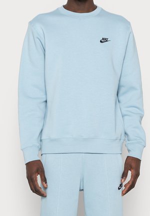 Homme portant un sweat-shirt Nike bleu clair et un pantalon de survêtement assorti avec des logos Nike noirs sur la poitrine et la cuisse.