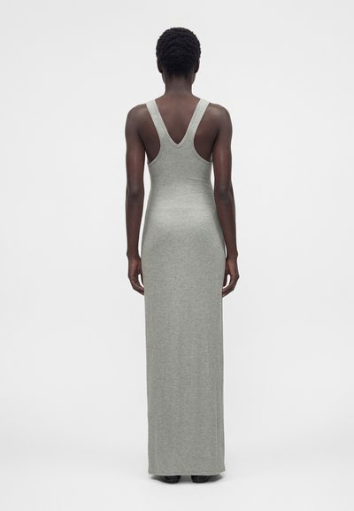 Γυναίκα με κοντά σγουρά μαλλιά που φοράει ένα αμάνικο, ανοιχτό γκρι, εφαρμοστό φόρεμα μέχρι το πάτωμα, στάζει με την πλάτη σε ένα απλό φόντο.