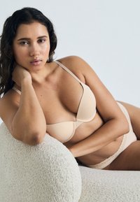 Nude lingerie set met een gewatteerde bh met een gladde afwerking en verstelbare banden, gecombineerd met bijpassende slipjes met kantafwerking, tegen een lichte achtergrond.