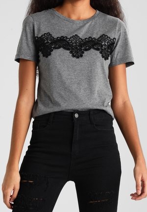 Vrouw draagt een grijs t-shirt met zwarte bloemenkant over de borst en zwarte, gescheurde high-waisted jeans.