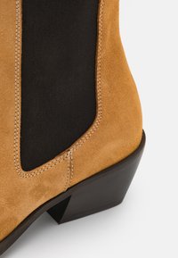 Tan mockasin med svart elastisk sidopanel, en staplad klack och kontrasterande vitt stygn längs sömmarna.