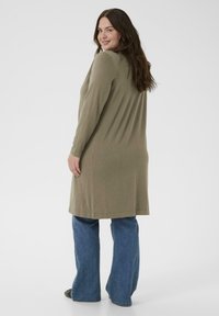 Kvinde der smiler over skuldren iført lang olivengrøn cardigan, blå jeans og sorte sko, stående mod en hvid baggrund.