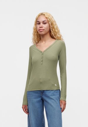 Giovane donna con lunghi capelli biondi ricci che indossa una maglietta verde oliva a maniche lunghe e jeans blu, in piedi davanti a uno sfondo semplice.