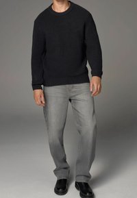 Homme portant un pull côtelé sombre, un jean gris et des bottes noires, se tenant contre un fond neutre avec une posture décontractée.