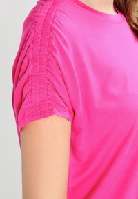 Detailní záběr osoby v jasně růžovém topu s krátkým rukávem, který má řasení podél ramenního švu.