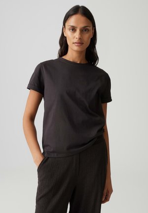Femme aux longs cheveux foncés, portant un t-shirt noir à manches courtes et un pantalon noir texturé, debout avec une main dans la poche sur un fond gris.