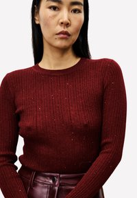 Rode geribbelde trui met lange mouwen, voorzien van subtiele metallic draden. Getailleerd ontwerp met een ronde halslijn, gecombineerd met een bordeauxrode leren riem.