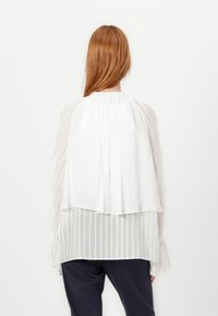 Blusa bianca a strati con design plissè, morbido al tatto e colletto arricciato. Maniche lunghe e fluide con lacci ai polsi.