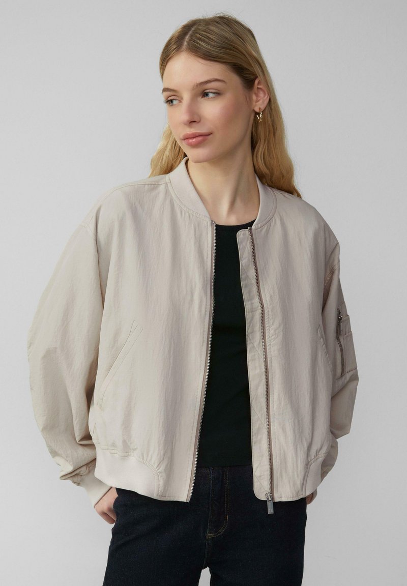 Lichtbeige bomberjack met ritssluiting, ribgebreide kraag en elastische manchet. Voorzien van zijzakken en een ritstas op de mouw.