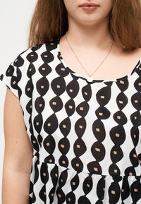 Γυναίκα που φοράει φόρεμα ασπρόμαυρο με σχέδιο, κοντά μανίκια και χρυσό κρεμαστό με στρογγυλό μενταγιόν, εμφανίζεται από τους ώμους μέχρι τη μέση.