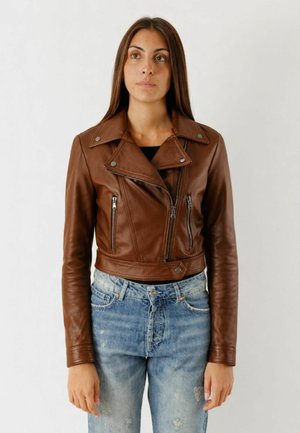 Donna con capelli lunghi che indossa una giacca di pelle marrone con cerniera e jeans chiari consumati, in piedi davanti a uno sfondo bianco.