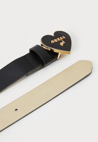 Une ceinture en cuir noir avec une boucle en forme de cœur dorée portant l'inscription "GUESS" en relief. L'autre sangle est en cuir beige, texture lisse.