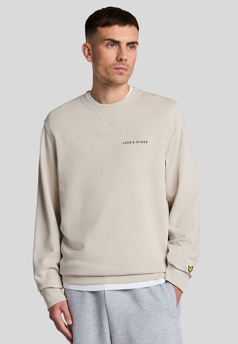 Sudadera beige con cuello redondo, mangas largas y un pequeño logo negro en el pecho. Decorada con un emblema amarillo en la manga.