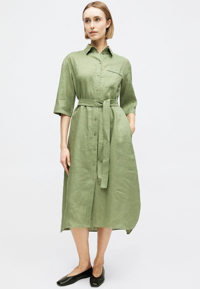 Γυναίκα που φοράει ανοιχτό πράσινο midi φόρεμα πουκάμισο με κουμπιά και ζώνη στη μέση, καθώς και μαύρα φλατ παπούτσια, στέκεται μπροστά σε έναν απλό λευκό φόντο.