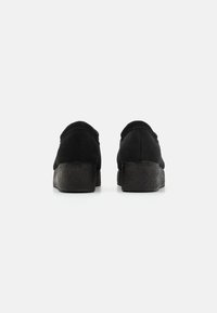 Juodi zomšiniai slip-on batai su tekstūrinėmis juodomis platforminėmis padais, apvaliais pirštais ir siuvinėjimu siūlėse.