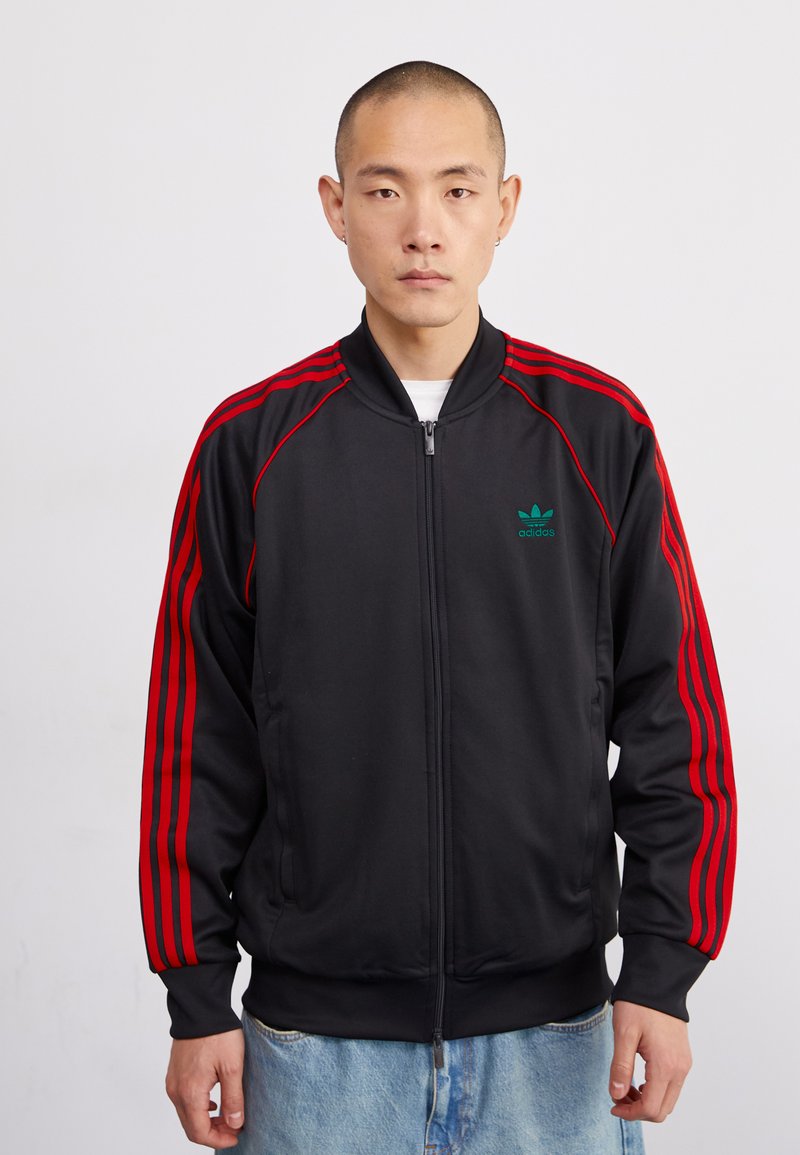 Černá zipová bunda se červenými trojpruhy, žebrovanými manžetami a límcem. Na levé straně hrudi se nachází malé zelené logo Adidas.