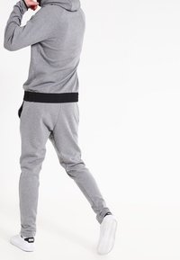 Šedá mikina s kapucí a tepláky s černým žebrovaným pasem. Tkanina je měkká a kalhoty se na kotnících zužují. Jsou vidět bílá sportovní obuv.