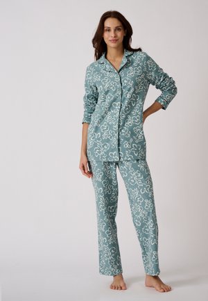 Femme debout pieds nus, vêtue d'un haut à manches longues boutonné à motif floral bleu sarcelle et d'un pantalon de pyjama assorti sur un fond uni.