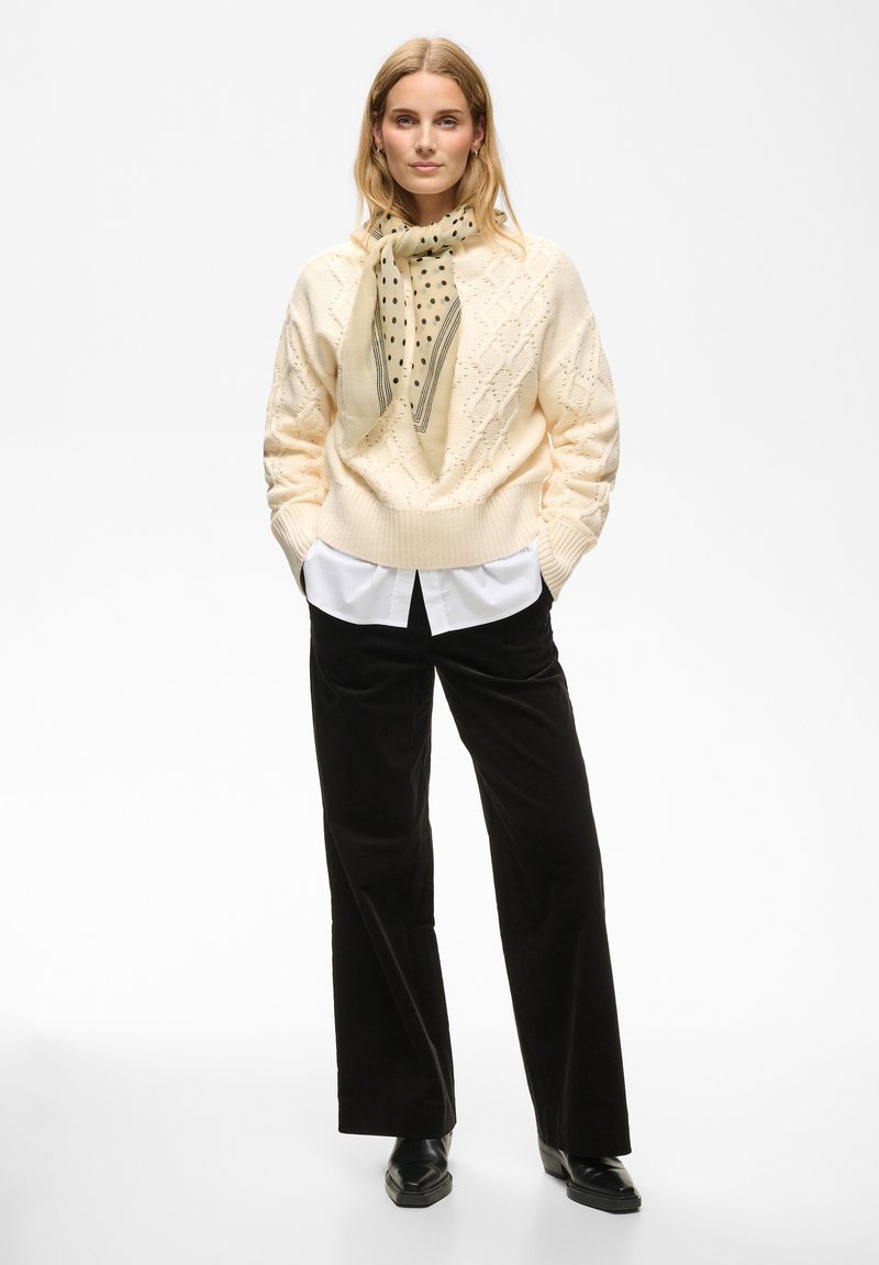 Femme debout, les mains dans les poches, portant un pull en maille torsadée crème, une écharpe à pois, une chemise blanche, un pantalon noir à jambes larges et des chaussures noires.