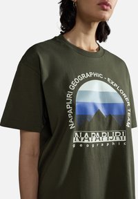 Camiseta de algodón color verde oliva con mangas cortas, que presenta un estampado gráfico de montañas y rayas en azul y blanco, además de un logo.