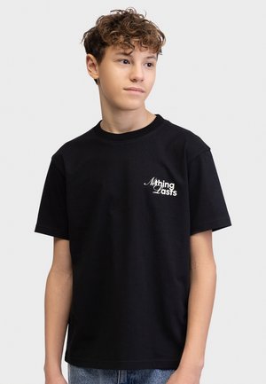 Έφηβος αγόρι με σγουρά μαλλιά που φοράει μαύρο T-shirt με λευκό κείμενο «Τίποτα δεν κρατάει» στο αριστερό μέρος του στήθους, στέκεται μπροστά σε απλό φωτεινό φόντο.