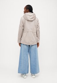 Veste à capuche beige clair avec une taille cintrée et des détails cousus, portée sur un pantalon en denim bleu clair à jambes larges, associé à des chaussures de sport.