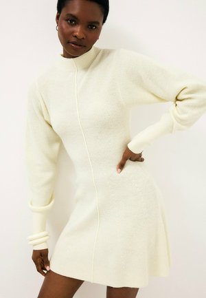 Woman wearing a cream ribbed knit long-sleeve dress with a high neckline, posing with one hand on her hip against a plain white background.
