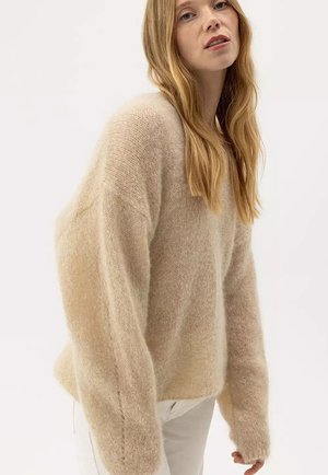 Mujer con cabello rubio largo que lleva un suéter beige suelto y peludo y pantalones blancos, posando sobre un fondo blanco liso.