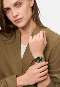 Goldene Armbanduhr mit quadratischem grünem Zifferblatt, das ein minimalistisches Design aufweist. Am Handgelenk getragen, zusammen mit einem braunen Blazer.
