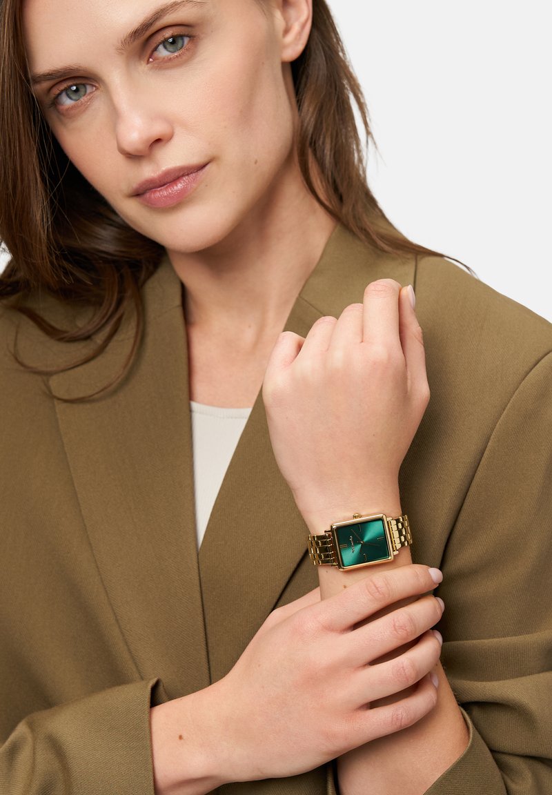 Goldene Armbanduhr mit quadratischem grünem Zifferblatt, das ein minimalistisches Design aufweist. Am Handgelenk getragen, zusammen mit einem braunen Blazer.