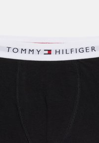 Μαύρα εσώρουχα με λευκή ελαστική ζώνη στη μέση που φέρει μαύρο κείμενο "Tommy Hilfiger" και το κόκκινο, λευκό και μπλε λογότυπο της μάρκας στο κέντρο.