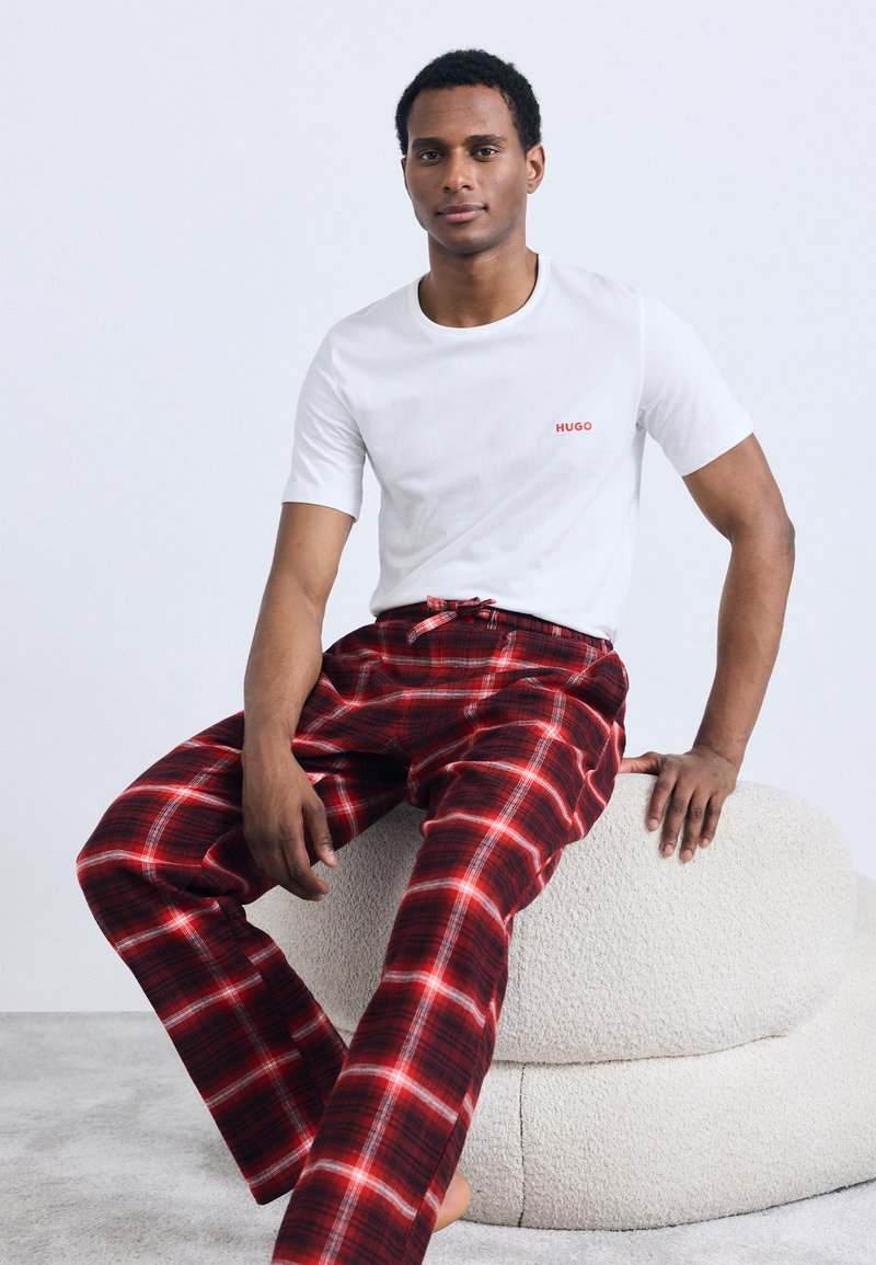 Camiseta blanca de manga corta con el logo rojo "HUGO", combinada con pantalones de pijama a cuadros rojos y negros, sentado en un cómodo otomán beige de forma redondeada.