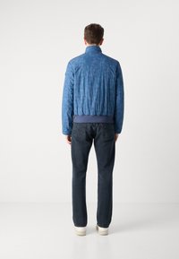Casaco bomber acolchoado azul com gola alta, cintura elástica e padrão tonal subtil, combinado com jeans escuros e sapatos claros.