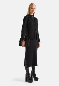 Černý upravený blazer s dvěma knoflíky, texturovaná tkanina; černá žebrovaná sukně; lesklé platformové boty po kolena; černá kožená kabelka s řetízkem.