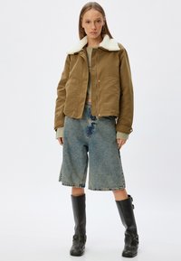 Brun jakke med hvid krave, grøn top, løse denim shorts, sorte knæhøje støvler med lommer og slidte kanter.