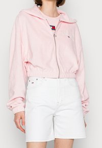 Lichtroze kortere zip-up hoodie gemaakt van zacht stof, met een logo op de borst, gecombineerd met witte denim shorts.