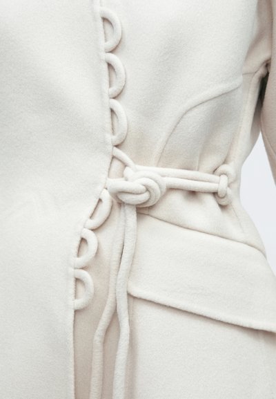 Λευκό παλτό από μιξ μαλλί με σχεδιασμό με βρόχους στο πλάι, λεπτομερή υφή υφάσματος και κλείσιμο με κόμπο στη μέση.