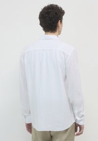 Camicia bianca a maniche lunghe con colletto, realizzata in tessuto leggero, caratterizzata da un yoke posteriore plissettato e polsini con un bottone.