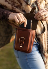 Bruine lederen telefoontas met een metalen sluiting en drukknoopjes. De tas staat rechtop, houdt stevig een zwarte telefoon vast en is bevestigd aan een riem.