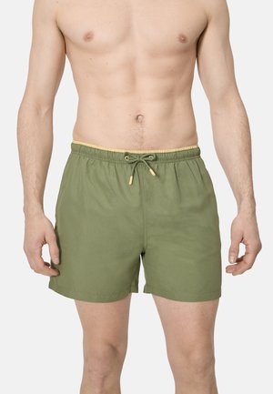 Torso y parte superior de las piernas de un hombre con bañador verde oliva con cordón amarillo y ribete en la cintura, de pie contra un fondo liso.