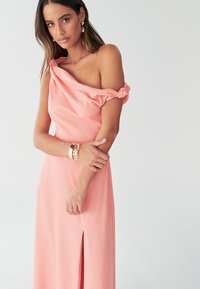 Šaty v broskvové barvě s odhalenými rameny, draped výstřihem a uzlovým detailem na rukávech. Obsahují rozparek až k stehnům pro zvýšení elegance. Hladká tkanina.