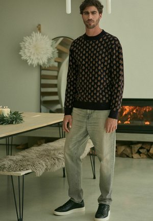 Maglione scuro con motivi ad albero, abbinato a jeans grigi chiari e sneaker nere. Ambientazione interna moderna con decorazioni minime.