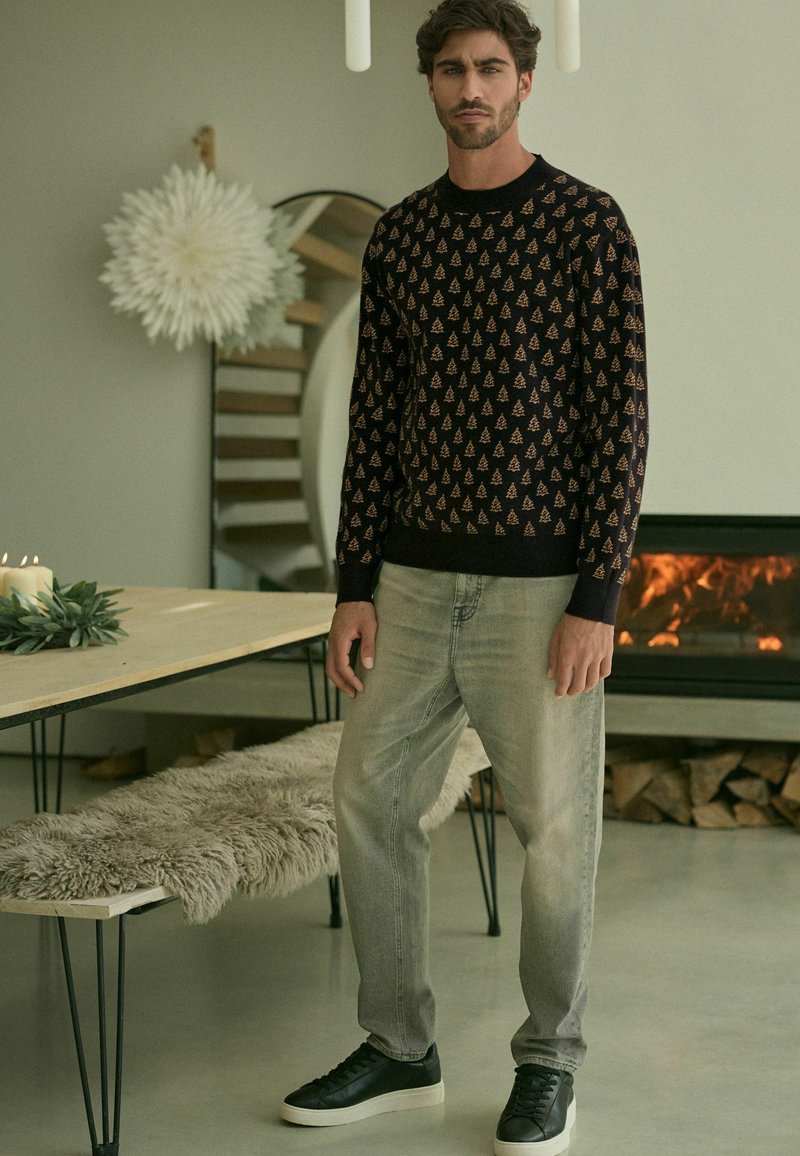 Maglione scuro con motivi ad albero, abbinato a jeans grigi chiari e sneaker nere. Ambientazione interna moderna con decorazioni minime.
