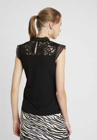 Zwarte top met korte kantmouwen, kantdetail op de rug en een knoopsluiting bij de hals. G gedragen met een rok in zebraprint.