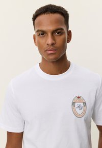 Λευκό βαμβακερό T-shirt με στρογγυλό λογότυπο σε κόκκινο, μπλε και κίτρινο χρώμα στο αριστερό στήθος. Μανίκια κοντά και κλασικός στρογγυλός λαιμός.